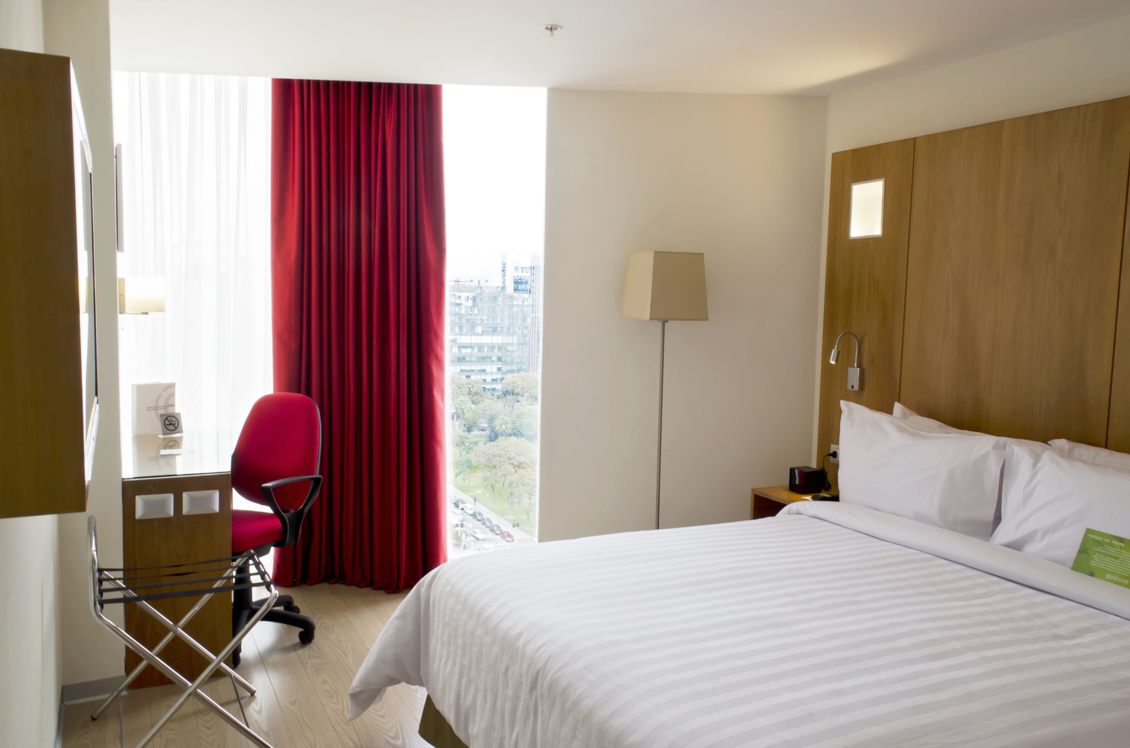 Ein Hotelzimmer mit Bett, Schreibtisch mit Stuhl, Lampe und Blick auf die Stadt durch große Fenster.