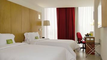 Habitación de hotel con dos camas, un escritorio con silla, una lámpara y una ventana con cortinas.