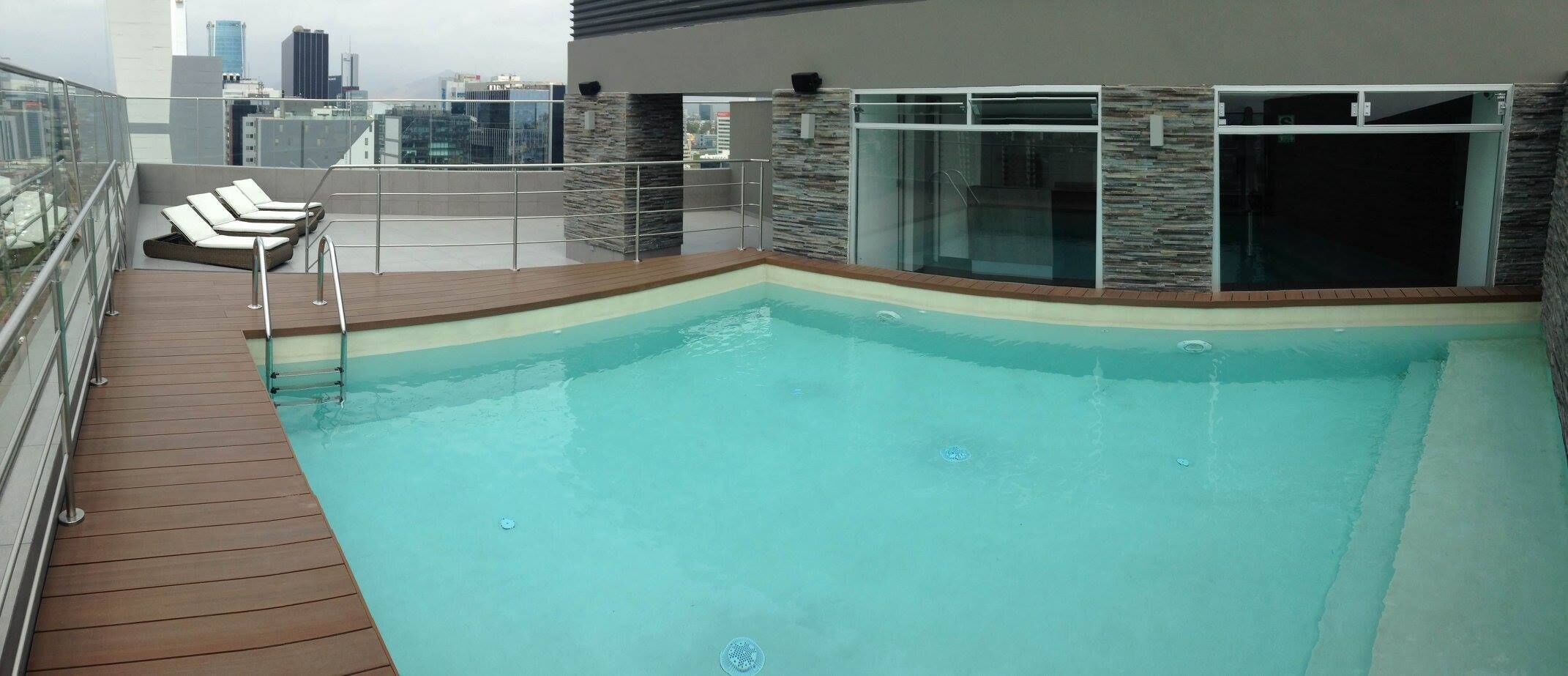 A rooftop pool with a wooden deck, glass balustrade, and lounge chairs.