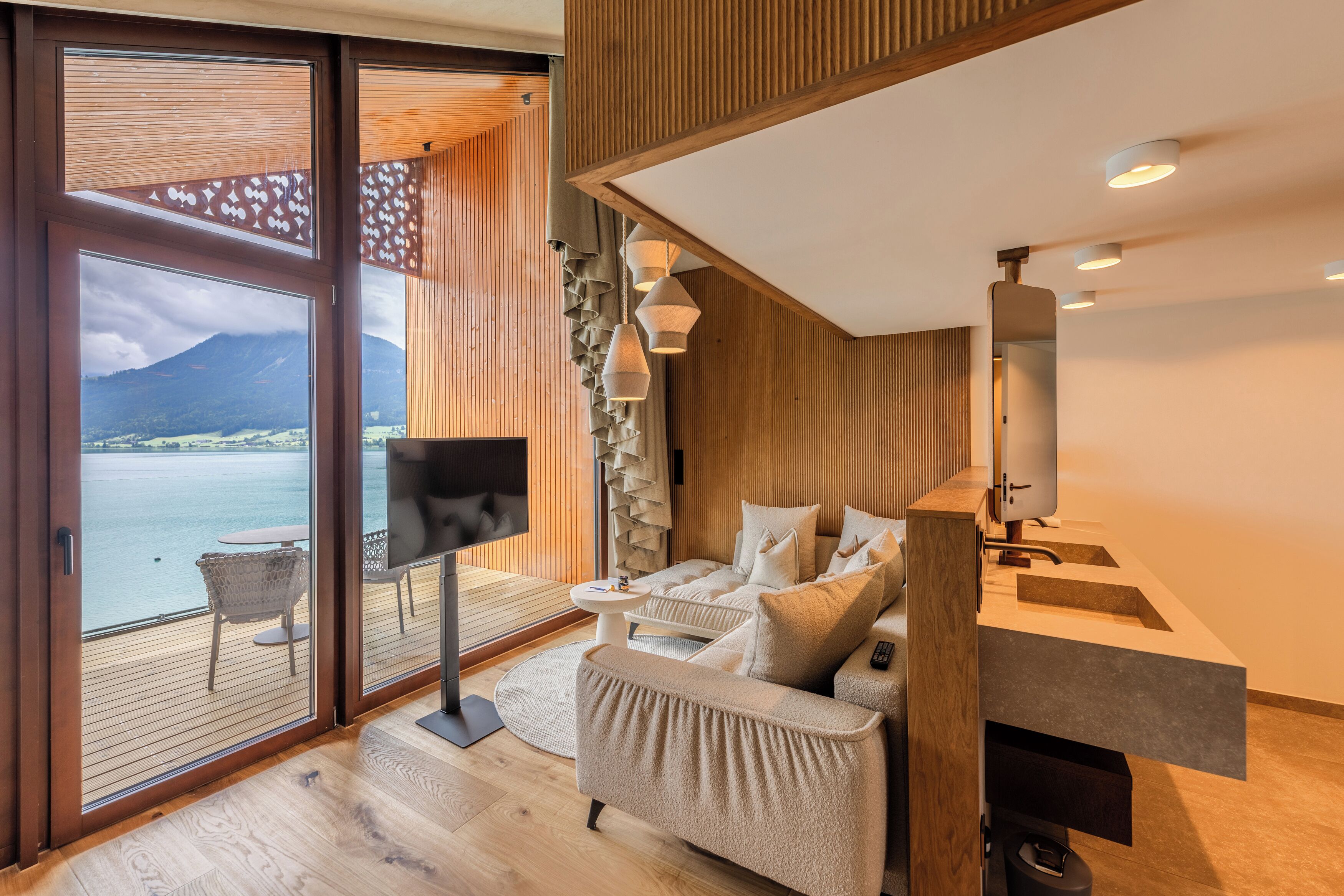 A modern interior with a balcony overlooking a body of water and mountains, a flat-screen TV, a sofa, and a bathroom with a mirror and sink.