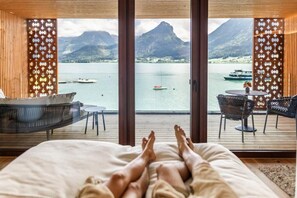 Una camera da letto con una grande finestra che si affaccia su un lago e montagne, un letto e due poltrone.