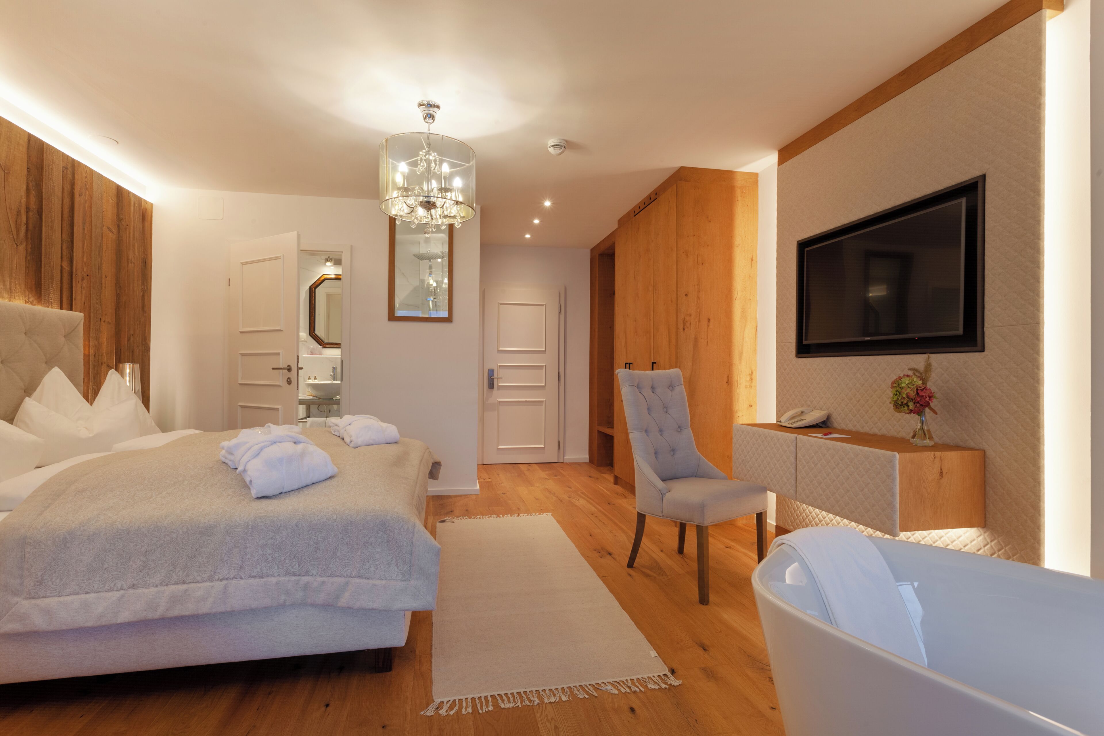 A modern bedroom with a large bed, a sitting area with a chair, a TV mounted on the wall, and a wooden wardrobe.
