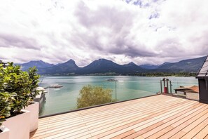 A wooden deck overlooking a calm lake with boats, surrounded by mountains and a cloudy sky. - Hotel Seevilla Wolfgangsee (Sankt Wolfgang im Salzkammergut)