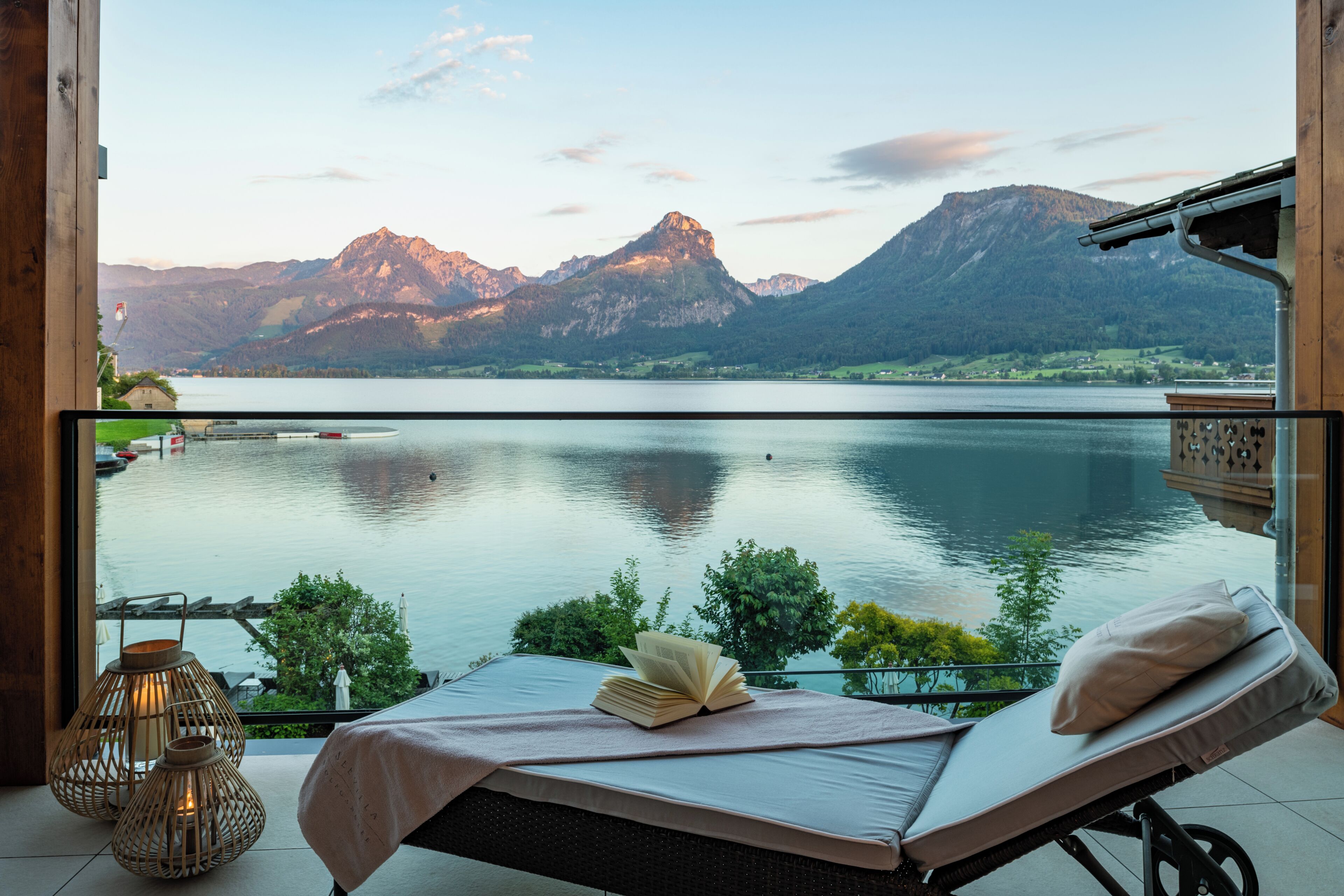 Un balcon avec vue sur un lac et des montagnes, une chaise longue et une table avec un livre et une bougie.