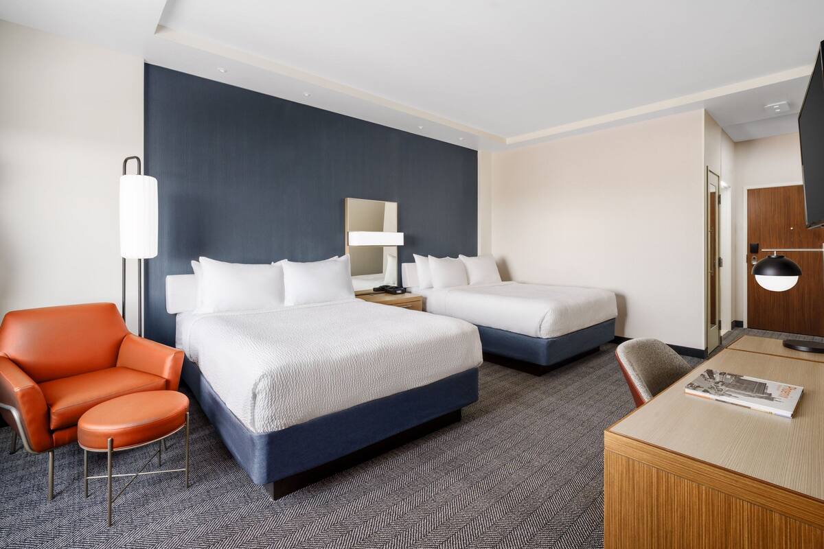 Quarto de hotel com duas camas, uma escrivaninha, uma cadeira e um espelho grande.