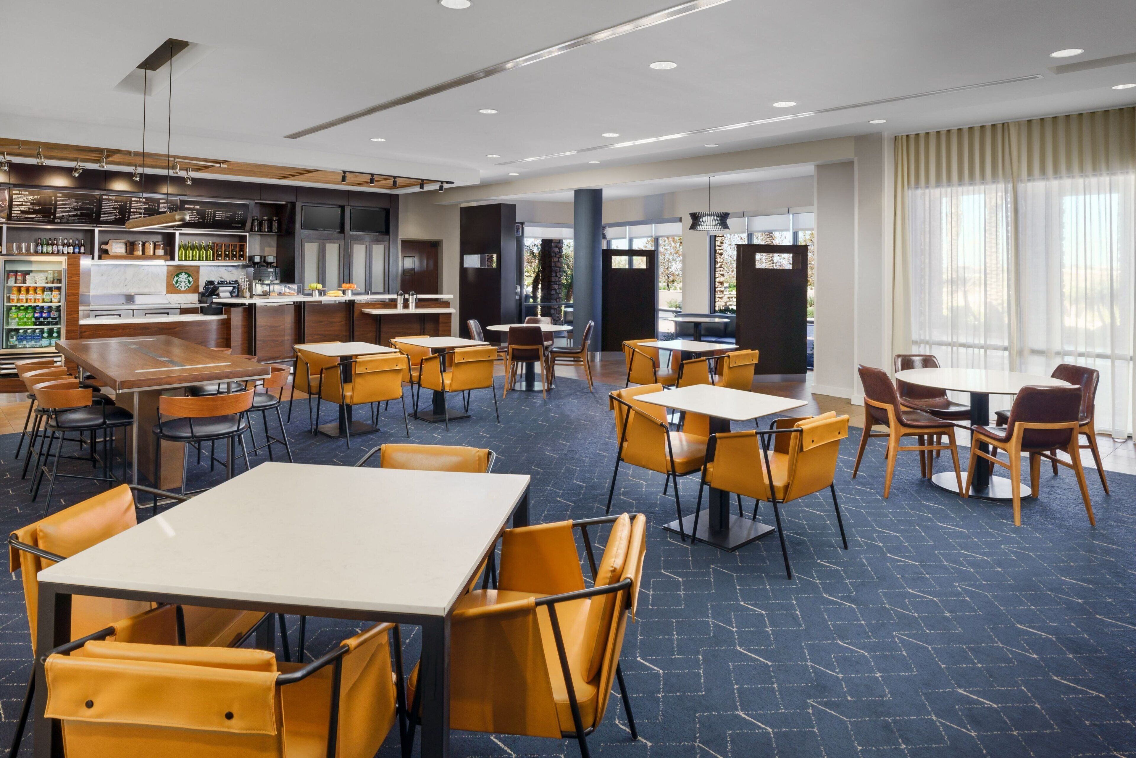 Un comedor moderno con varias mesas y sillas, una zona de bar con una máquina de café, y vistas al exterior a través de grandes ventanales.