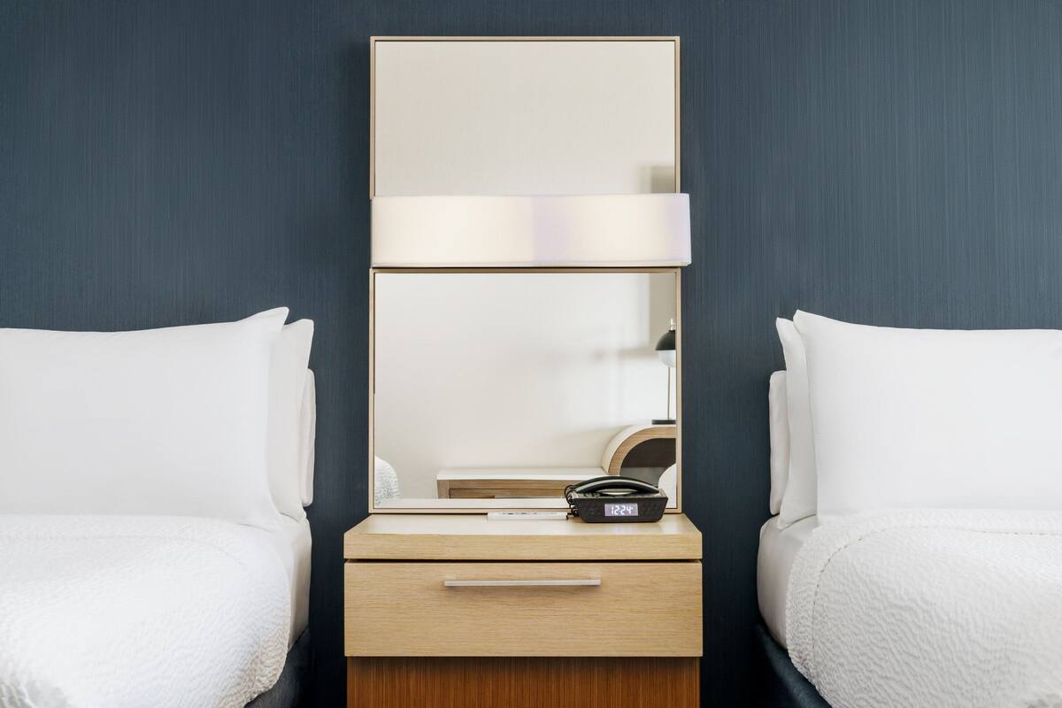 Quarto de hotel com duas camas de solteiro, mesa de cabeceira com espelho, luminária e relógio.
