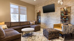 A modern living room with a sectional sofa, a round coffee table, and patterned ottomans. A flat-screen TV is mounted on the brick wall, and there is a large window providing natural light. - Drury Inn & Suites Louisville North (Louisville)