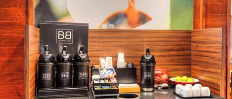 A coffee station with branded dispensers, a tray of snacks, and a bowl of fruit.