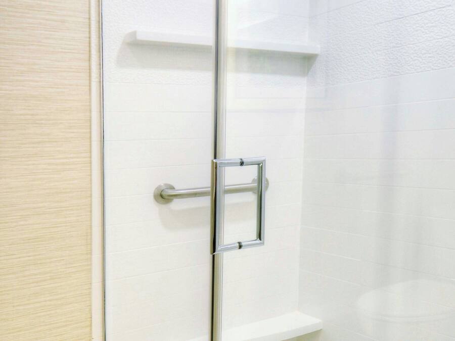 Una ducha con cerramiento de vidrio, pared de azulejos blancos, un asa metálico y una repisa.