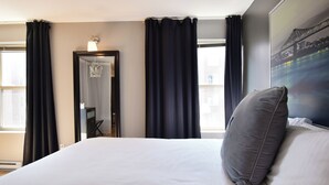 A hotel room with a large bed, dark curtains, a mirror, and a cityscape painting on the wall. - Lester Lofts by Bower Boutique Hotels (Moncton)