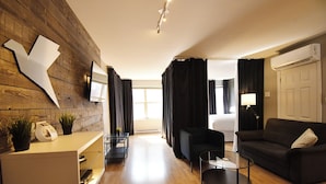 A modern hotel room with a wooden accent wall, a flat-screen TV, a sofa, a coffee table, and a bed with black curtains. - Lester Lofts by Bower Boutique Hotels (Moncton)