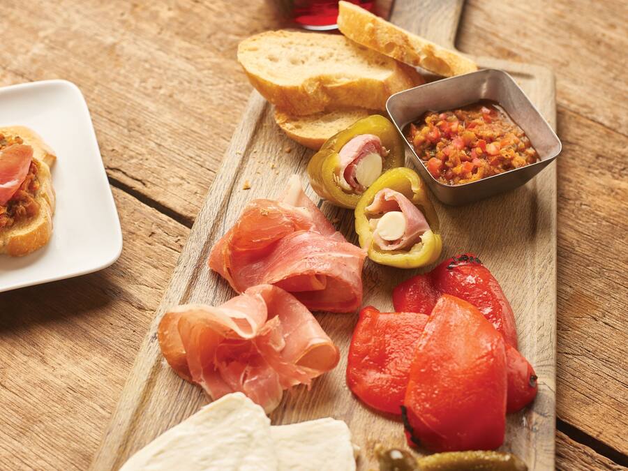 Tabla de madera con diversos alimentos, entre ellos tomates en rodajas, pepinillos y pan, acompañada de una copa de vino tinto.