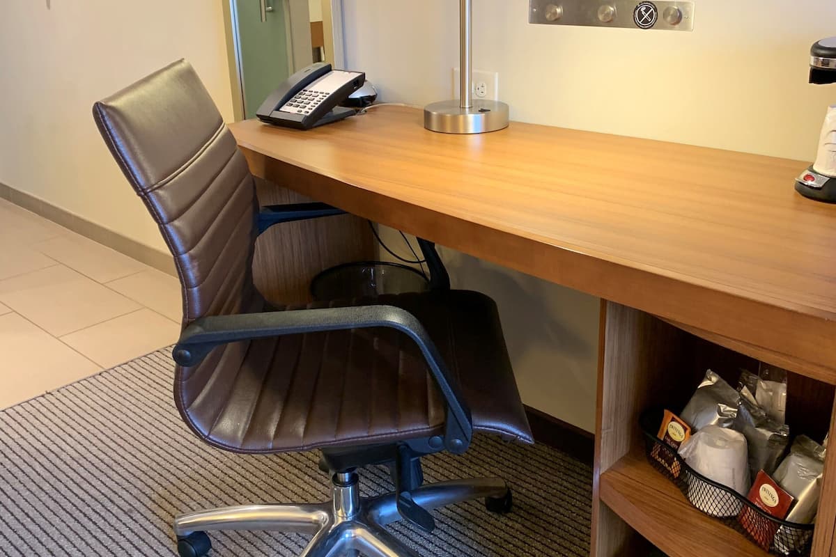 A hotel office with a desk, chair, phone, lamp, and a shelf with various items.