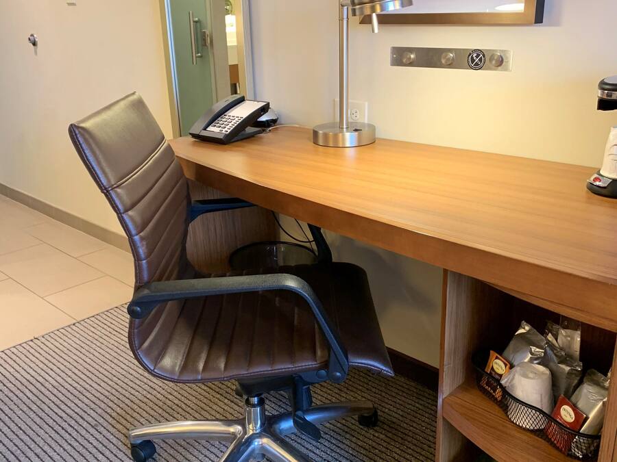 A hotel office with a desk, chair, phone, lamp, and a shelf with various items.