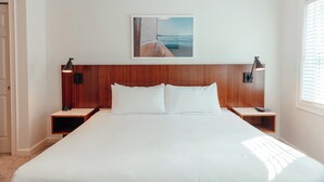 Ein ordentlich eingerichtetes Hotelzimmer mit einem großen Bett, einem Holzkopfstück, zwei Nachttischen mit Lampen, einem gerahmten Bild an der Wand und einem Fenster mit Jalousien.
