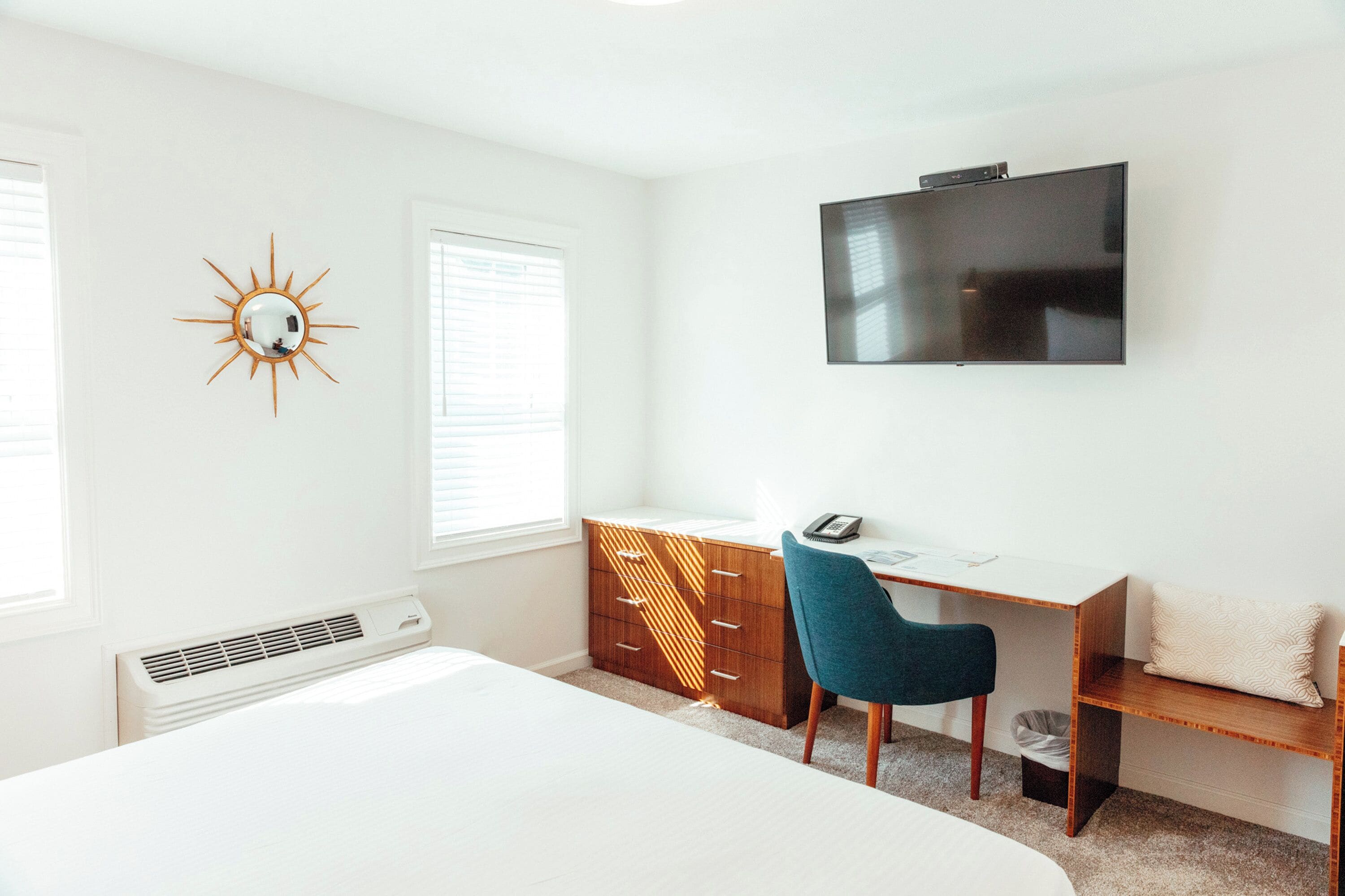 Une chambre à coucher avec un lit, un bureau avec une chaise, une télévision et un miroir décoratif en forme de soleil accroché au mur.
