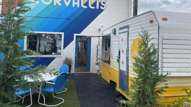 A hotel named 'hotel CORVALLIS' with a colorful mural, a small outdoor seating area with blue chairs, and a vintage camper parked in front.