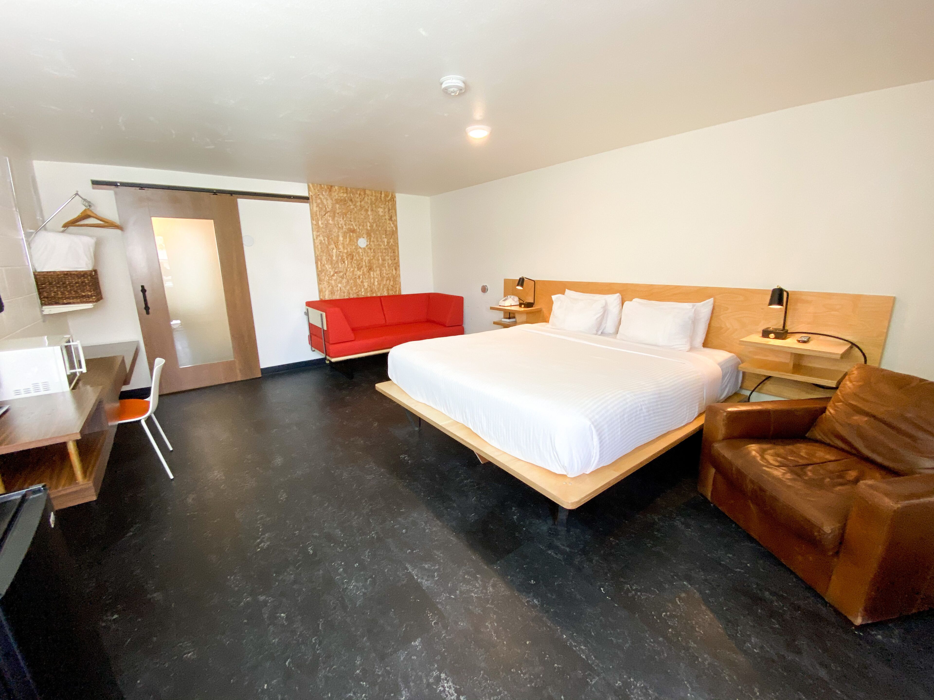 A modern hotel room with a large bed, a desk, a red sofa, and a leather armchair.