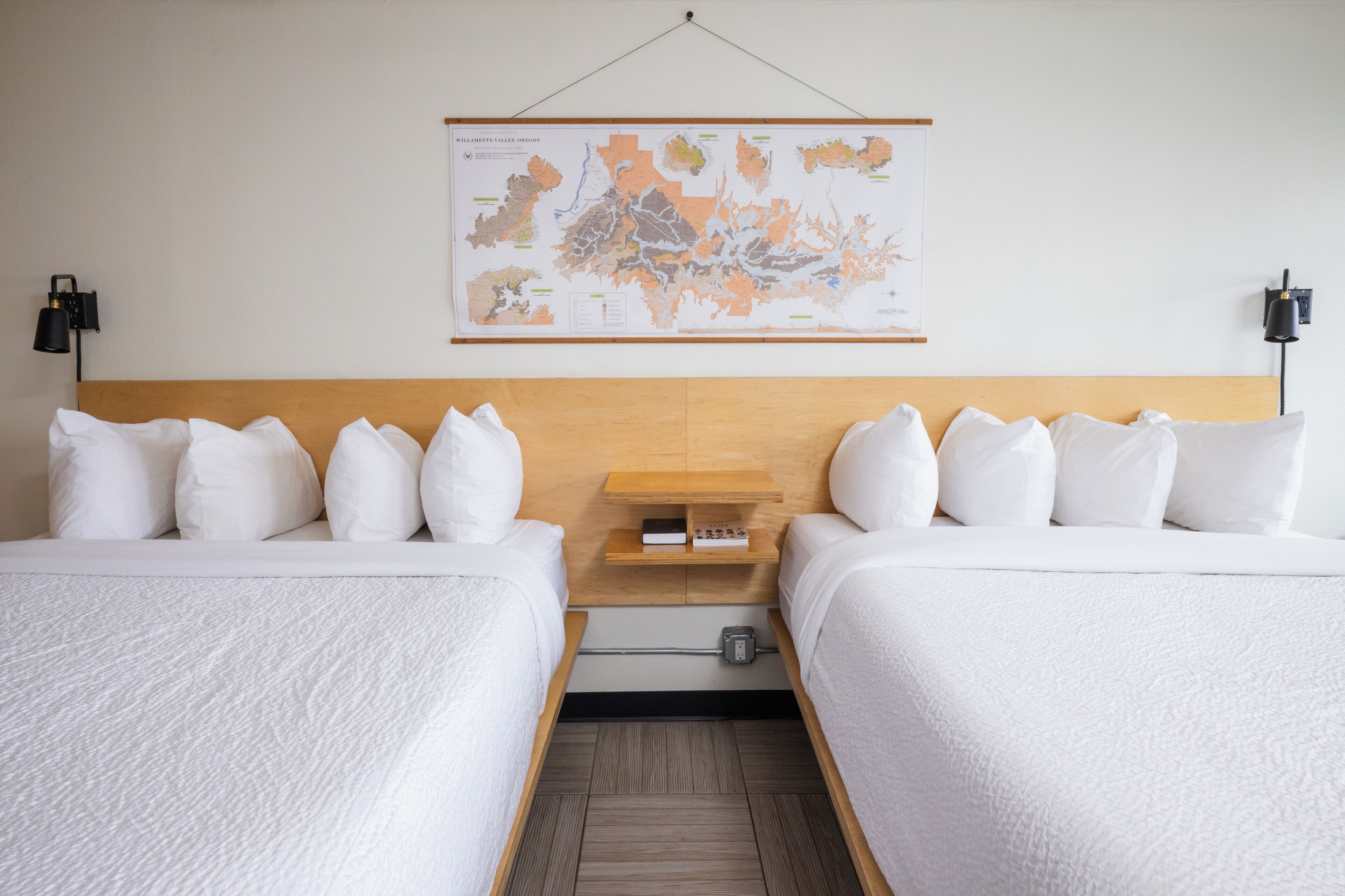 A hotel room with two beds, a wooden headboard, a map on the wall, and a small table with a book and a lamp.