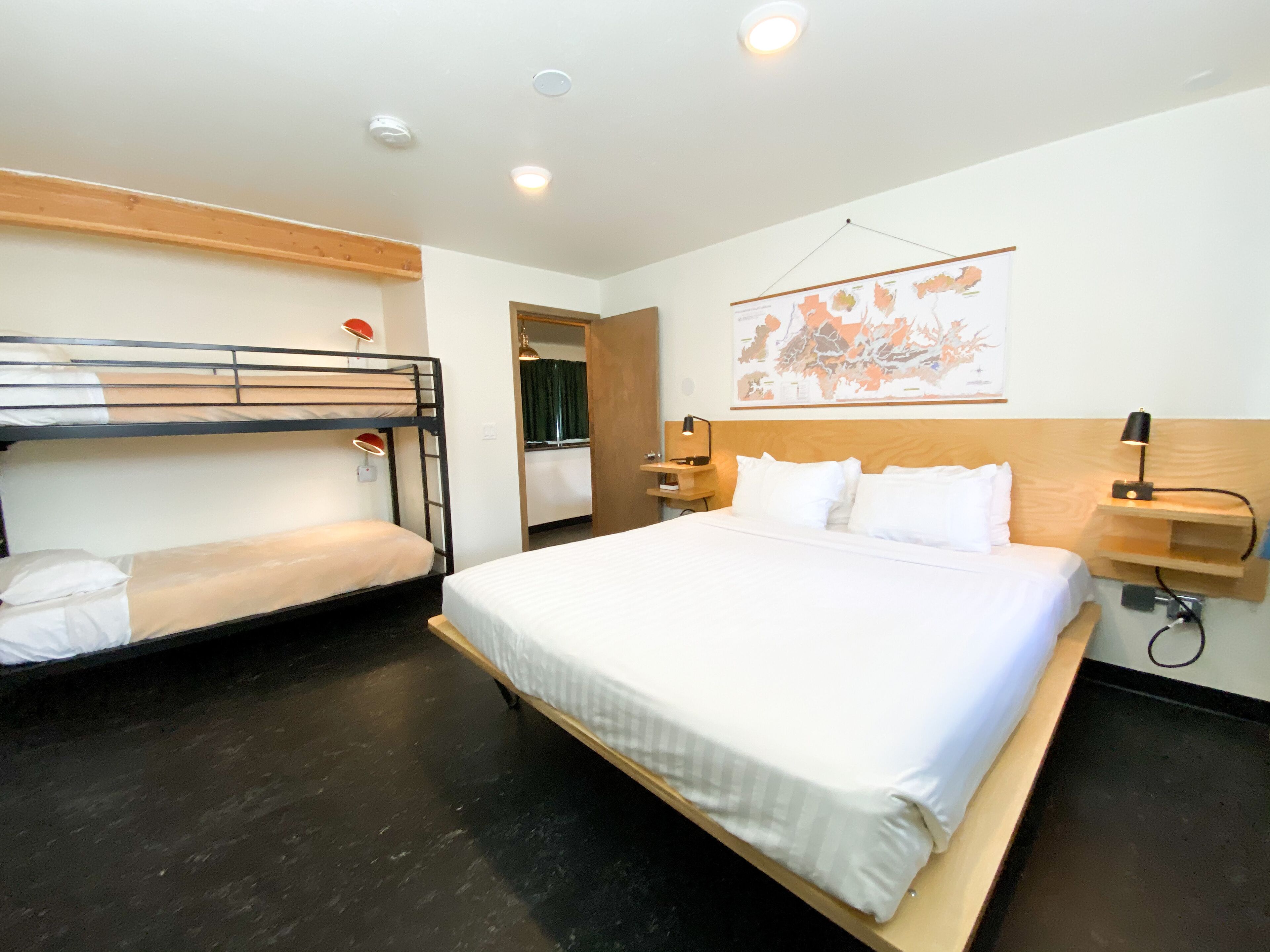 A hotel room with a bunk bed, a single bed, a wooden headboard, a map on the wall, and a nightstand with a lamp.