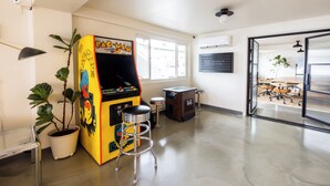 A spacious room with a large window, a potted plant, a colorful arcade machine, and a drum set visible through an open door.