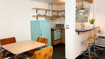 A compact kitchen with a blue refrigerator, a microwave, and a sink. There is a dining area with a wooden table and orange chairs.