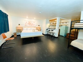 Habitación de hotel compacta con cama, escritorio, silla, litera y kitchenette.
