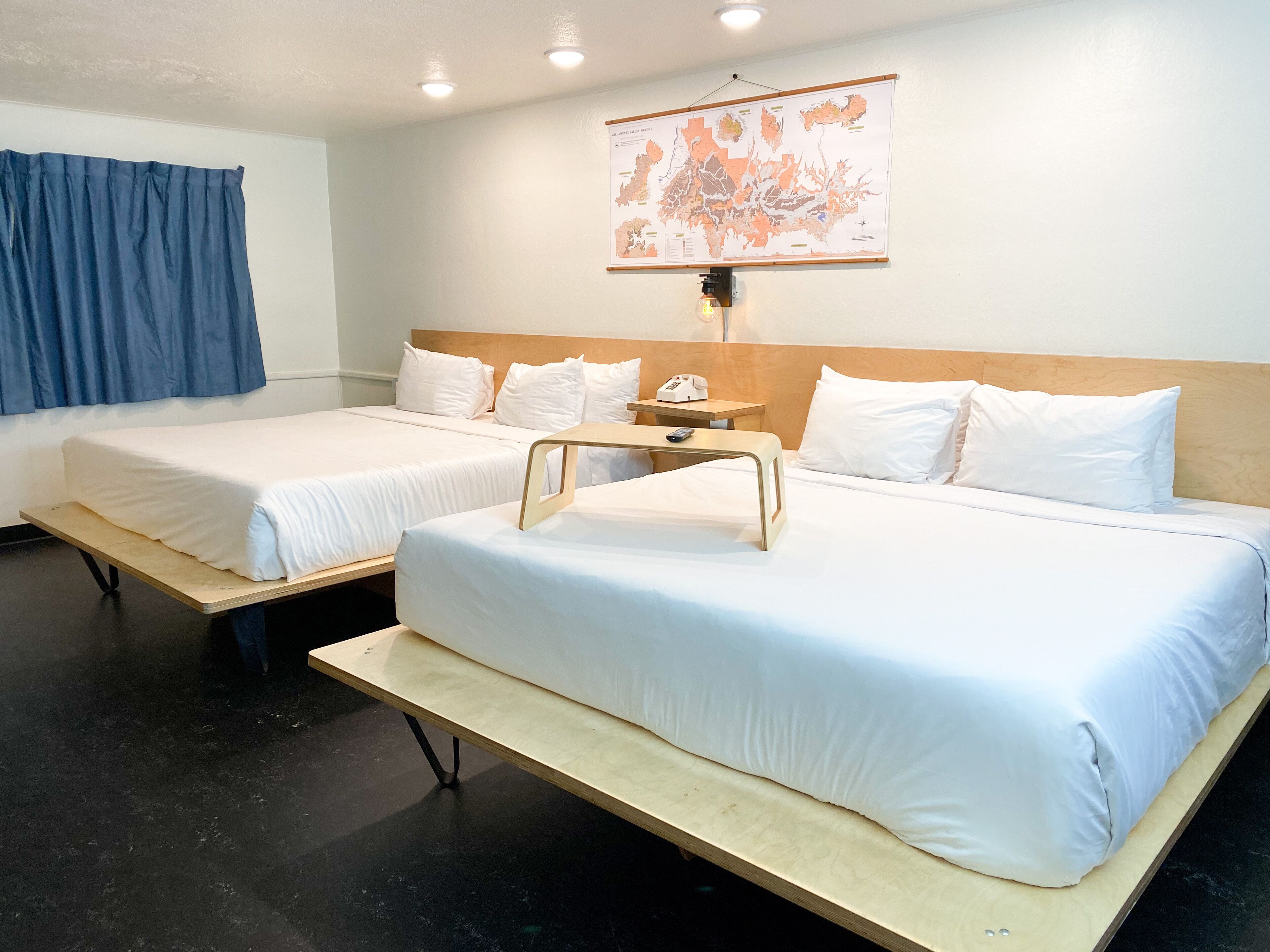 A hotel room with two single beds, a wooden headboard, a small bedside table with a lamp, and a map on the wall.
