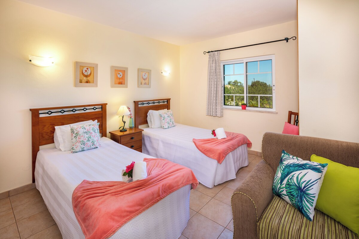 A hotel room with two beds, a sofa, and a window with a view of greenery.