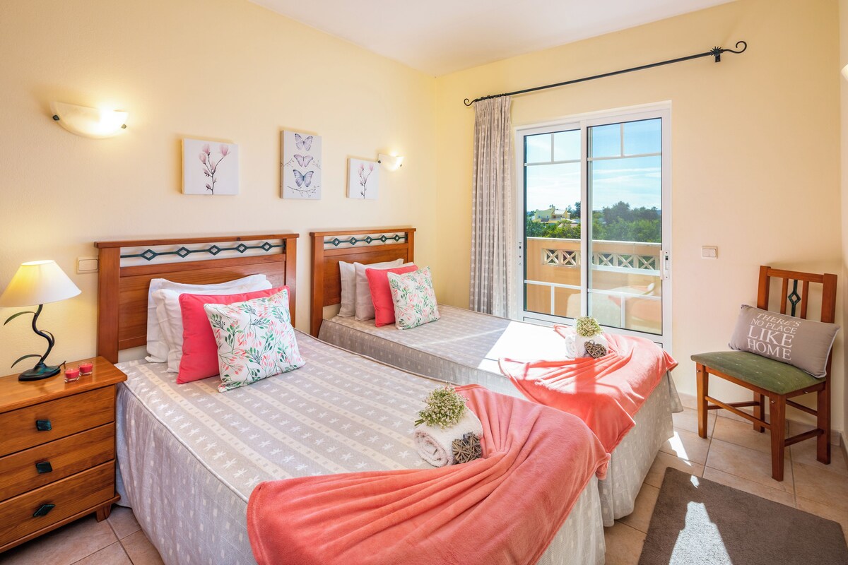 A bedroom with two beds, a nightstand, a chair, and a balcony with a view.