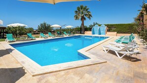 A swimming pool with lounge chairs and umbrellas, surrounded by greenery and a clear sky.