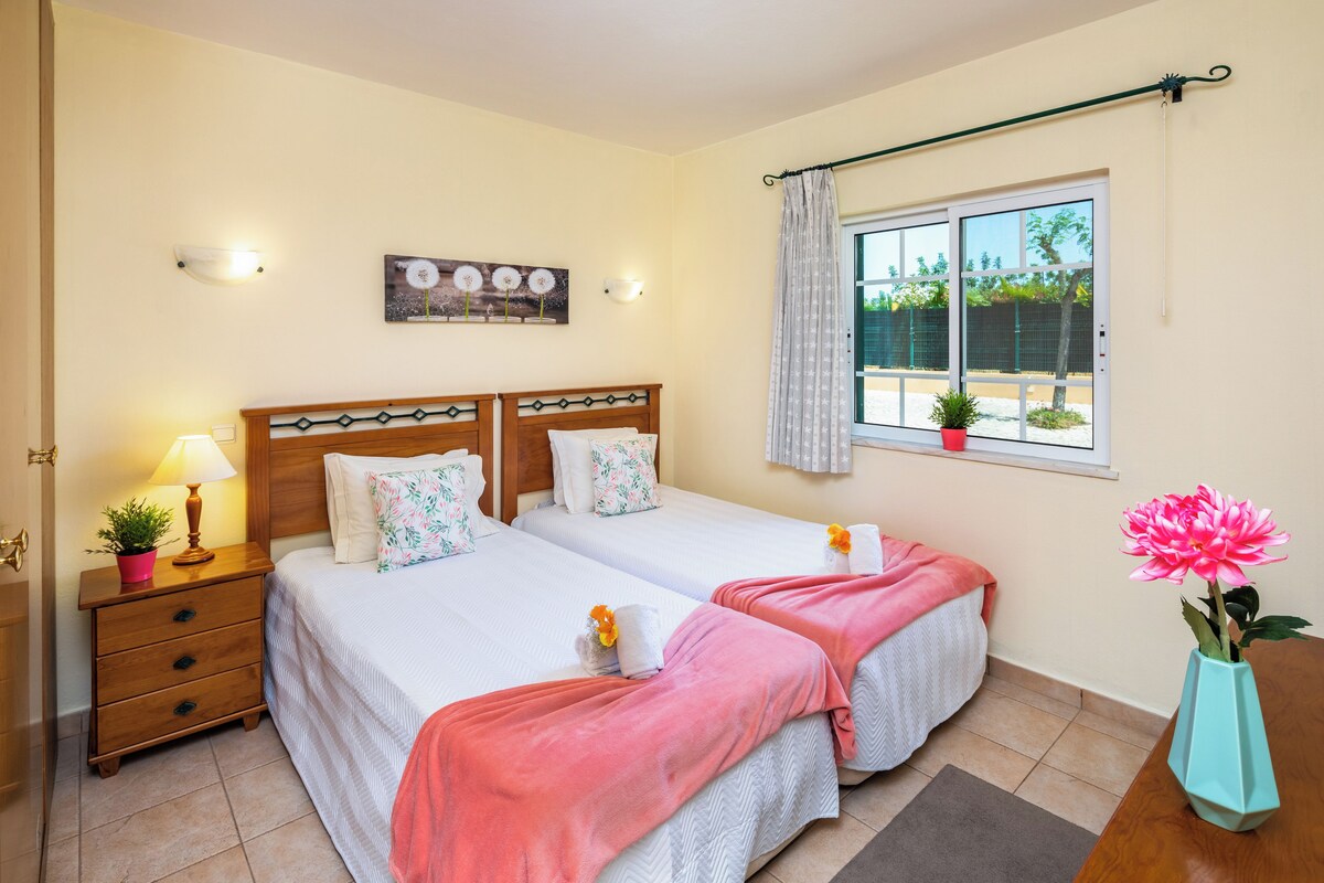 A double room with two beds, a bedside table with a lamp, a window with a view, and a potted plant.