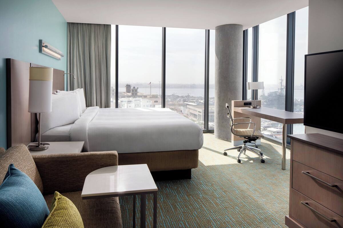 A hotel room with a large bed, a desk, a chair, a TV, and a view of the city.
