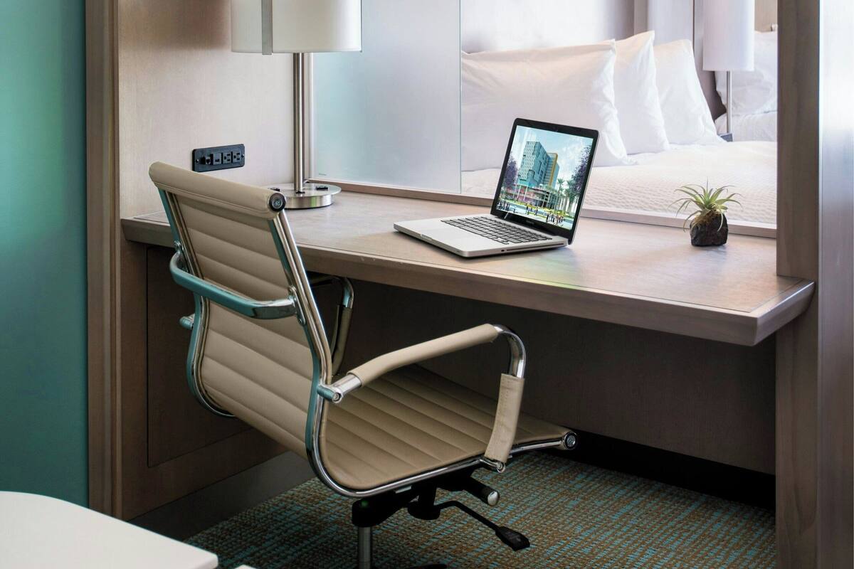 A hotel room with a desk, chair, laptop, bed, and a small plant.