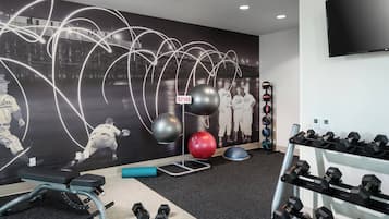 Uma academia com equipamentos de exercícios, um mural na parede e uma televisão fixada na parede.