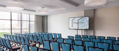 Un salón de conferencias con filas de sillas azules, una pantalla grande que muestra 'SPRINGHILL SUITES' y ventanas amplias con vistas a edificios.