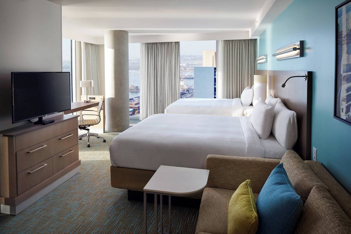 A hotel room with a large bed, a TV, a desk, and a view of the city.