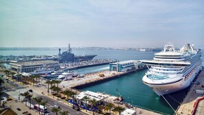 Een cruiseschip in de haven, met een grote scheepswerf en militaire schepen op de achtergrond.