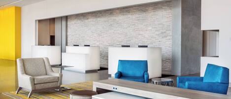 A modern lobby with a reception desk, two seating areas with blue and beige chairs, a patterned rug, and a brick accent wall.