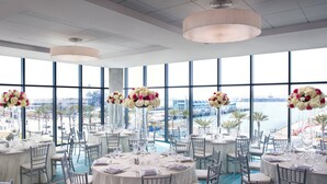 Een eetgedeelte met ronde tafels gedekt voor een formeel evenement, grote ramen met uitzicht op de stad en elegante bloemdecoraties.