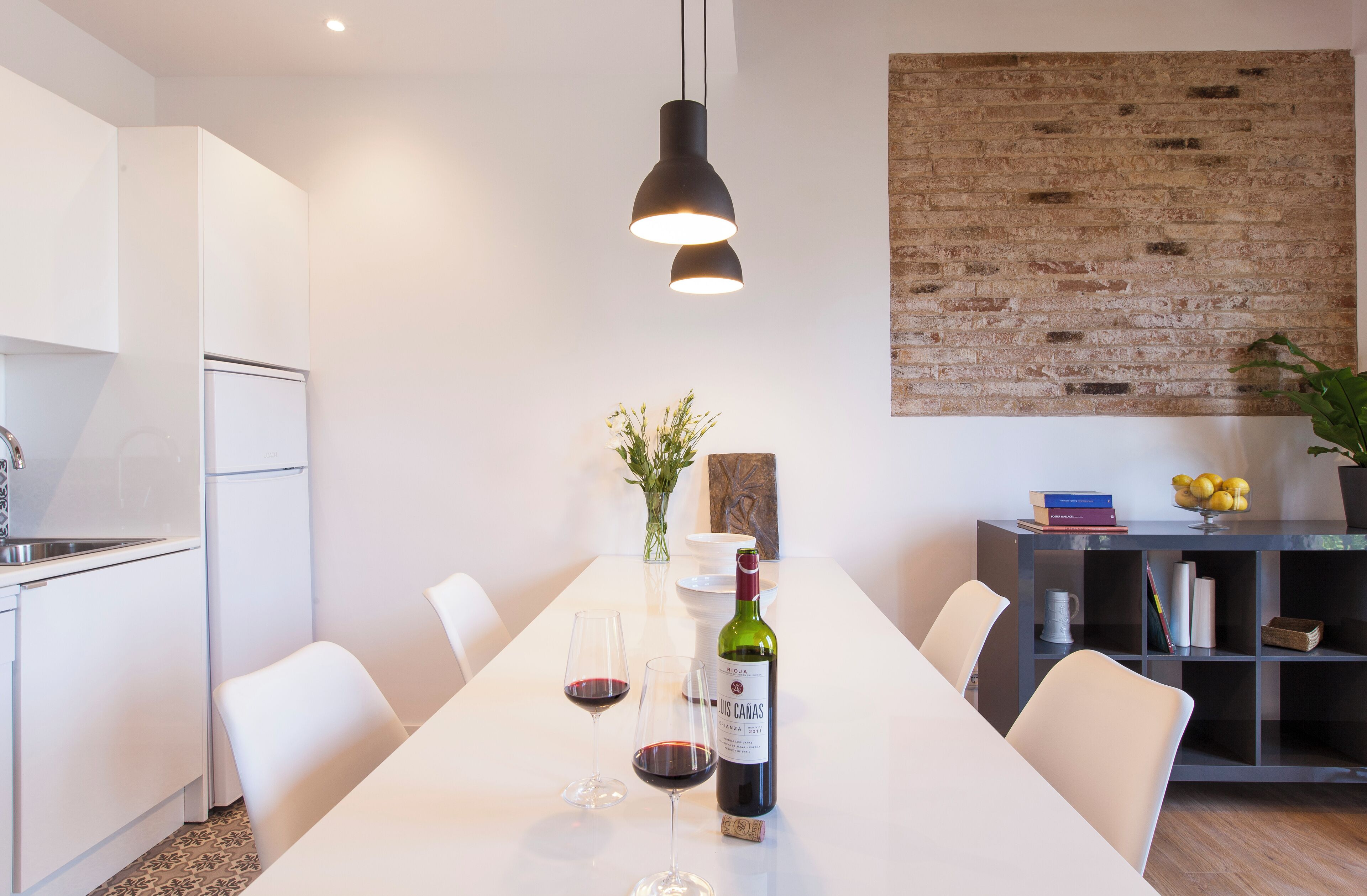 A modern kitchen with white cabinets, a dining table with chairs, a hanging light fixture, and a brick accent wall.