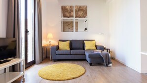 A modern living room with a grey sofa, yellow cushions, a round yellow rug, a flat-screen TV on a white stand, and a large window with curtains. - Aspasios Charming Flats (Barcelona)