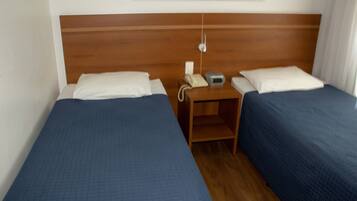 Quarto de hotel com duas camas de solteiro, cada uma com cabeceira de madeira, mesa de cabeceira com telefone e relógio digital, e uma parede branca lisa.