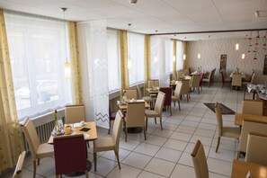 A spacious restaurant with multiple tables and chairs, large windows with curtains, and hanging pendant lights.