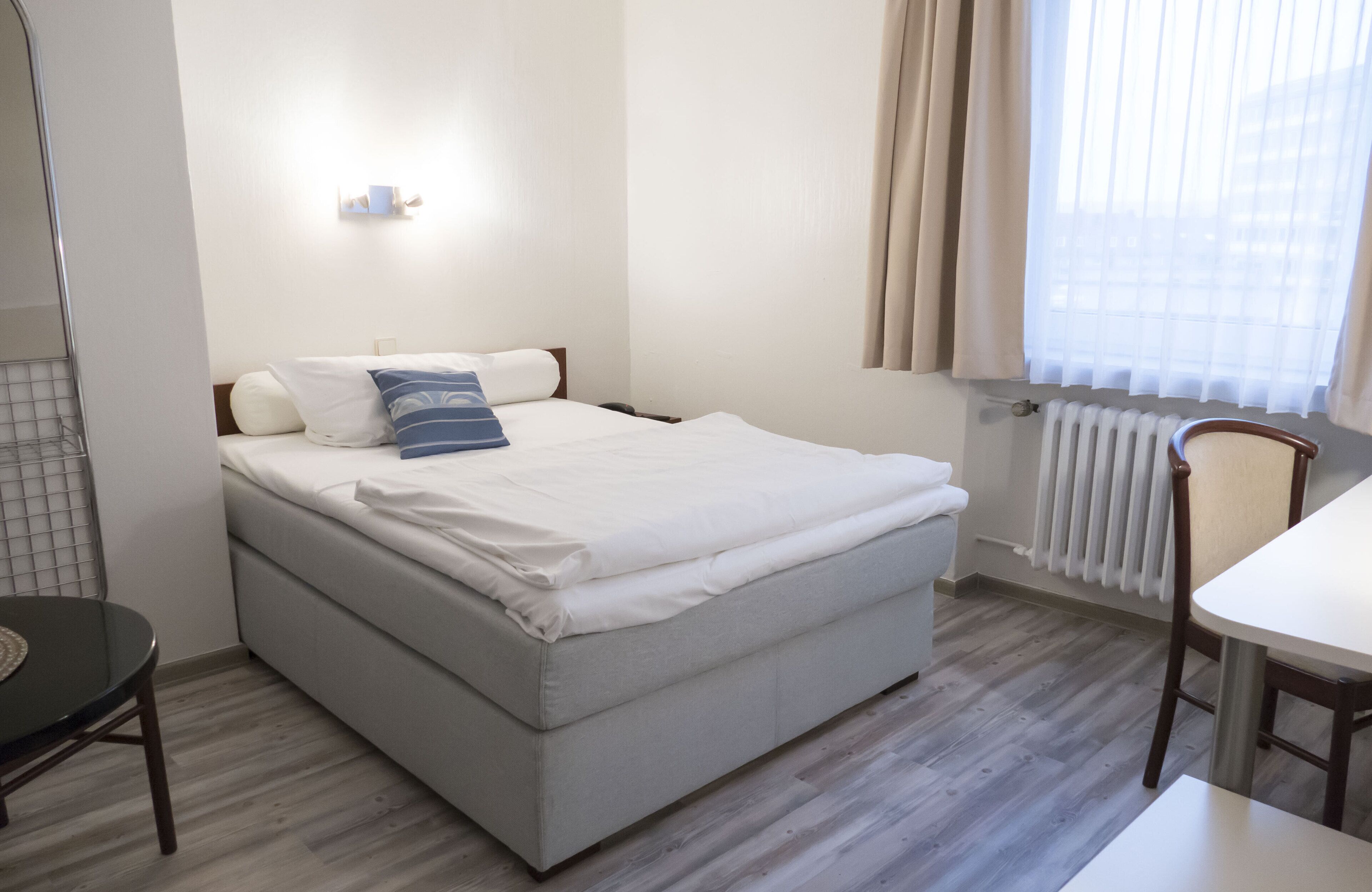 A single bed with a white mattress and two pillows, a small table, a chair, a wall-mounted light, and a window with curtains.