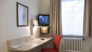 A hotel room with a desk, a television, a red chair, a framed picture on the wall, and a window with curtains. - Hotel Excelsior (Kassel)