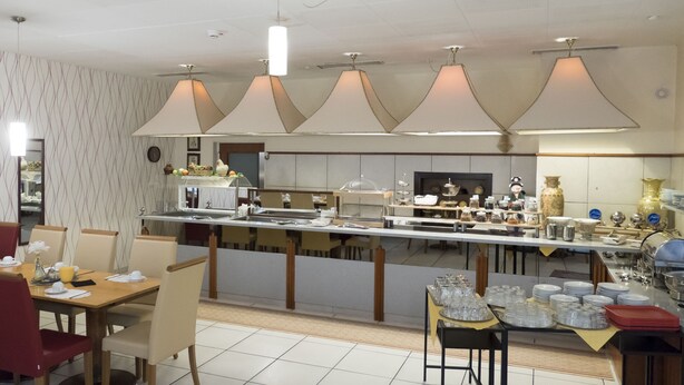 A restaurant with a long counter, multiple tables, and chairs. There are hanging lamps above the counter and a clock on the wall. - Hotel Excelsior (Kassel)