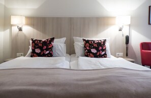 A hotel room with two beds, each with white linens and floral-patterned pillows. There are bedside lamps and a red chair.