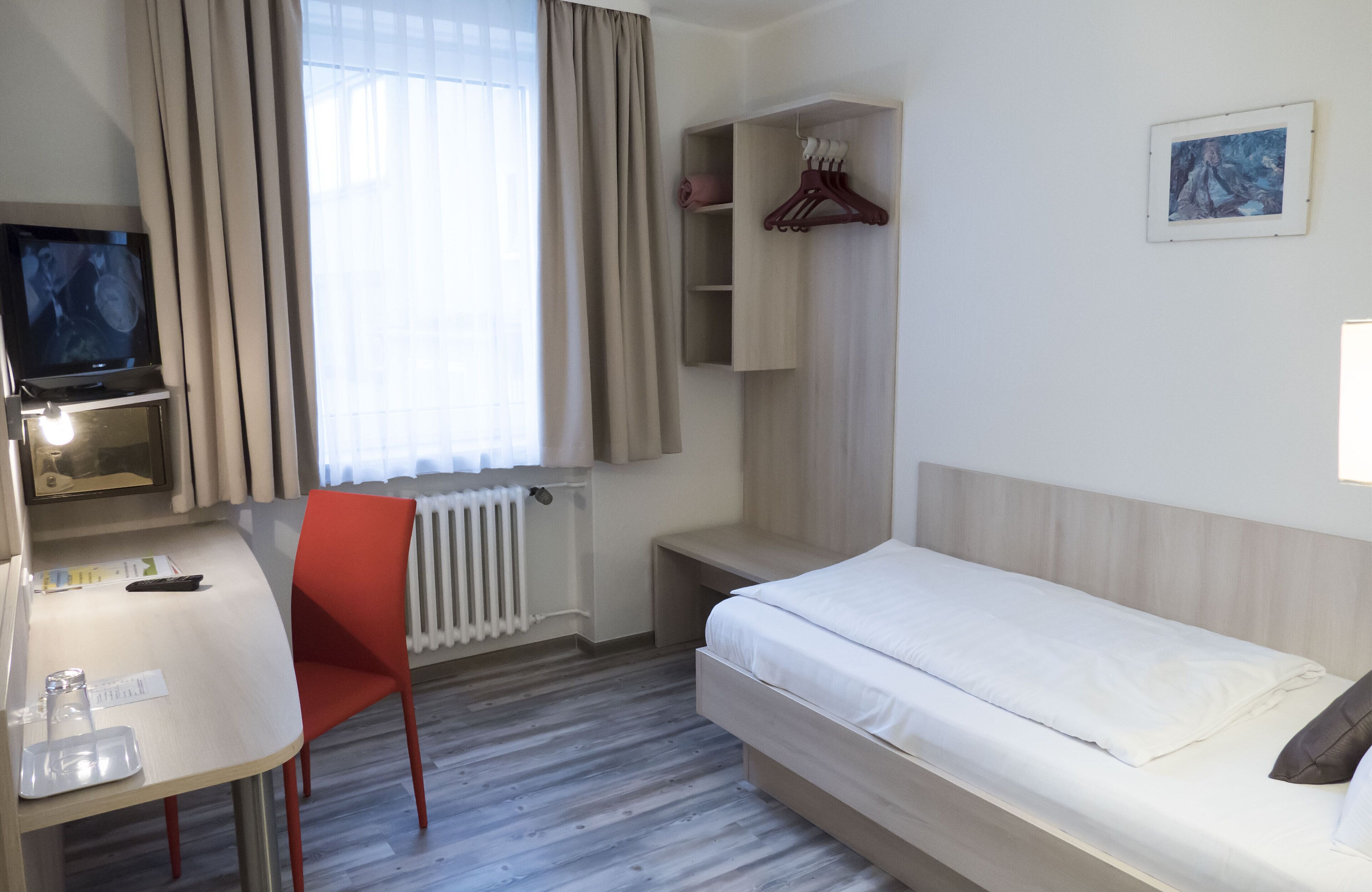 A hotel room with a bed, a desk with a chair, a TV, a radiator, and a wardrobe.